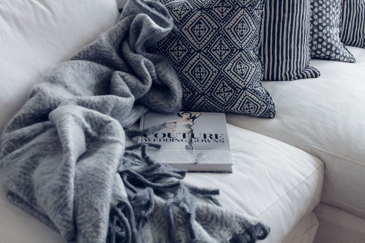 A stylish living room featuring a white sofa with patterned pillows, a blanket, and a fashion book.