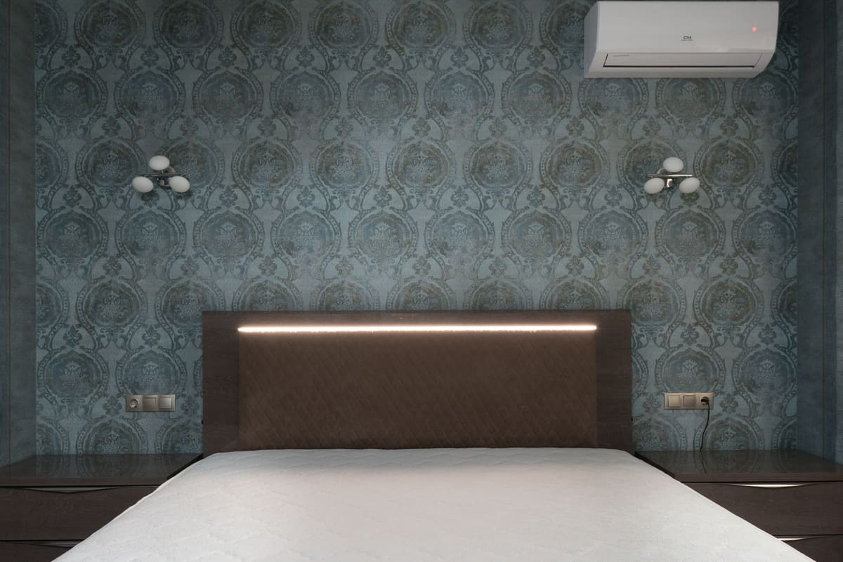 White mattress on bed with brown headboard placed between bedside tables at wall with ornamental wallpaper and decorative white sconces carpet head